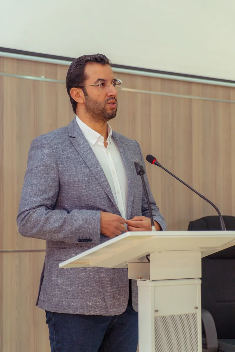 Dr CHARRASS Abdelmajid debout à un pupitre et parlant dans deux microphones.