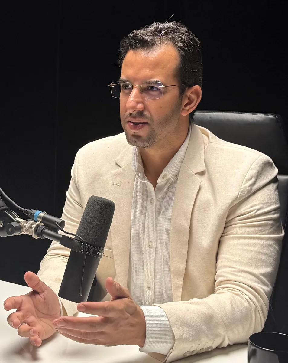 Dr CHARRASS Abdelmajid en veste claire devant un microphone noir, mains ouvertes.