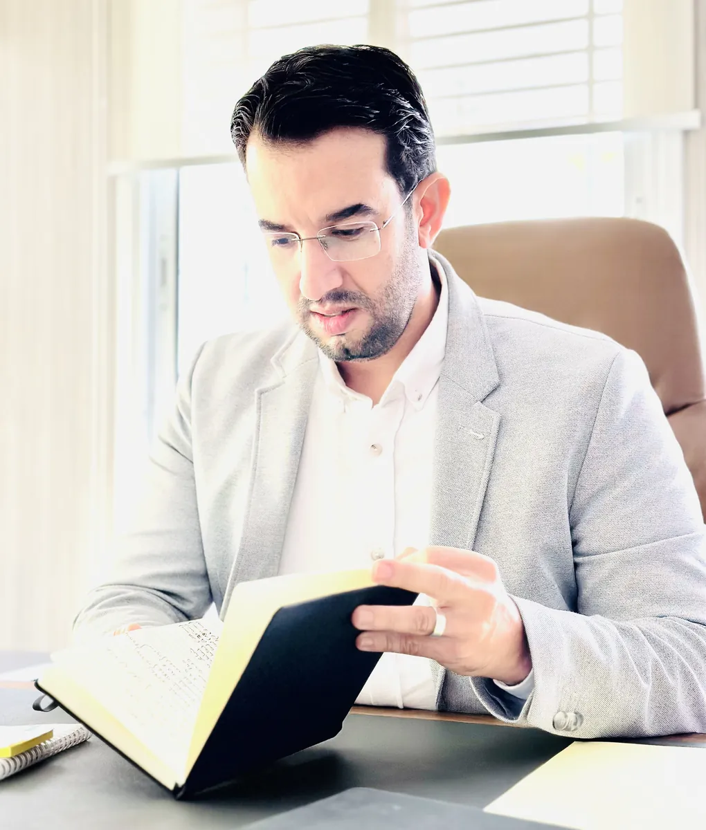 Dr CHARRASS Abdelmajid en veste grise lisant un livre ouvert a son bureau.