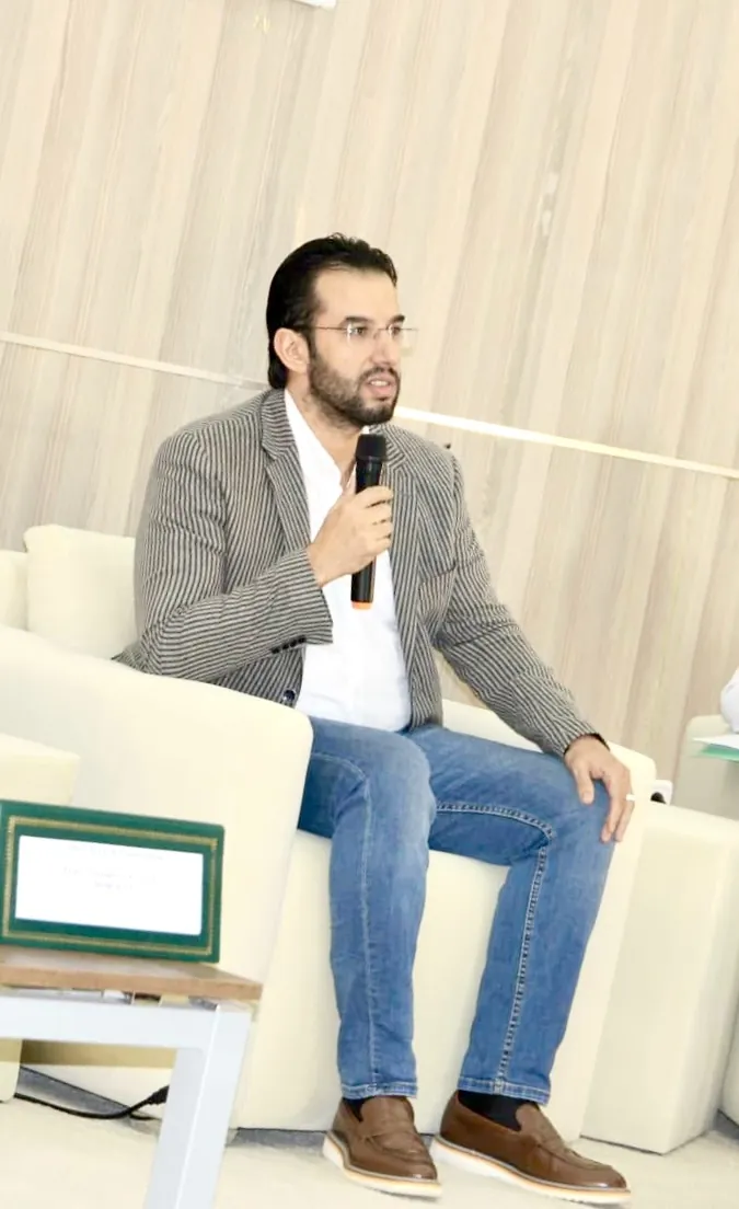 Dr CHARRASS Abdelmajid en costume gris assis dans un fauteuil lors d’un panel.